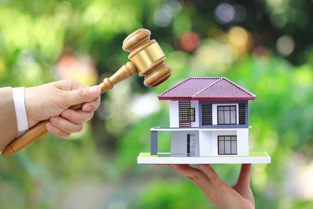 Casa all’asta: mano femminile che tiene un martelletto accanto a una casa in miniatura su sfondo bianco, concetto di avvocato immobiliare e proprietà